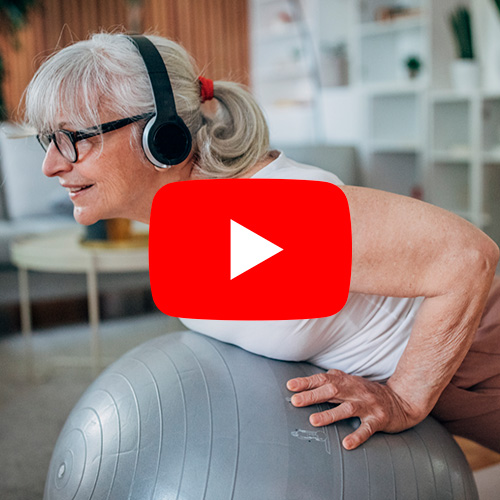 Mulher idosa com fones de ouvido praticando exercício físico sobre bola de pilates em casa, associando estilo de vida saudável ao equilíbrio hormonal.