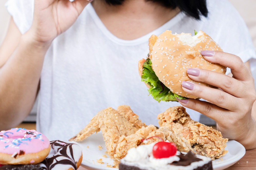 Pessoa segura hambúrguer e frango frito, com mesa cheia de doces como donuts e bolo, sugerindo compulsão alimentar.