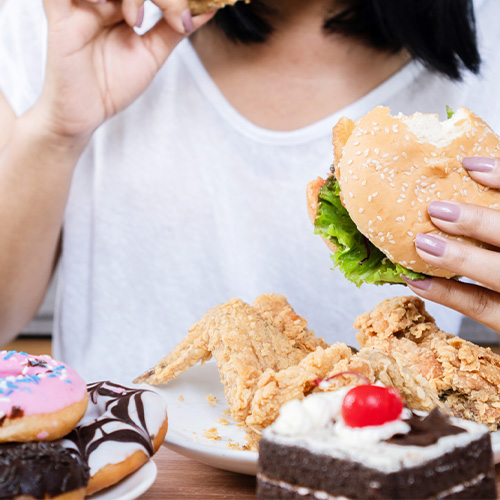 Pessoa segura hambúrguer e frango frito, com mesa cheia de doces como donuts e bolo, sugerindo compulsão alimentar.