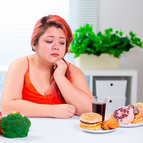 Mulher com expressão preocupada observa uma mesa com alimentos ultraprocessados e saudáveis, refletindo sobre escolhas alimentares.