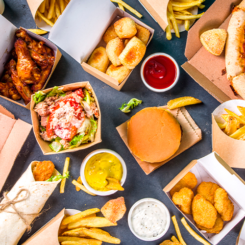 Variedade de fast foods sobre a mesa, com hambúrguer, batatas fritas, nuggets, molhos e sanduíches em embalagens descartáveis.