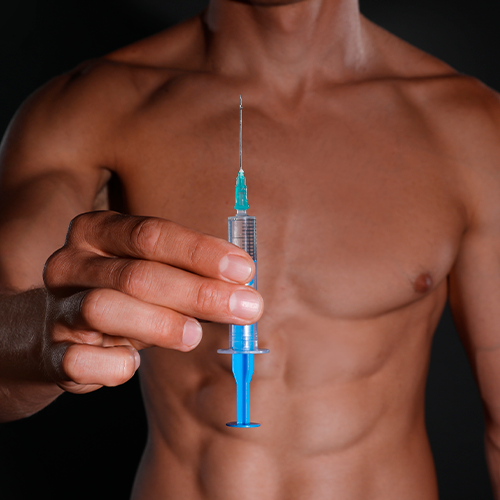 Homem sem camisa segura uma seringa com líquido azul à frente do corpo, em fundo escuro.