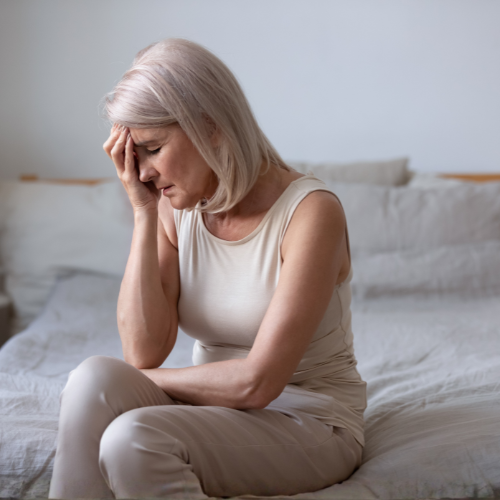 Mulher sentada na cama com a mão na testa, expressão de cansaço ou mal-estar em ambiente doméstico.