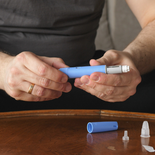 Pessoa segurando caneta injetável para medicação, com peças do aplicador sobre mesa de madeira.