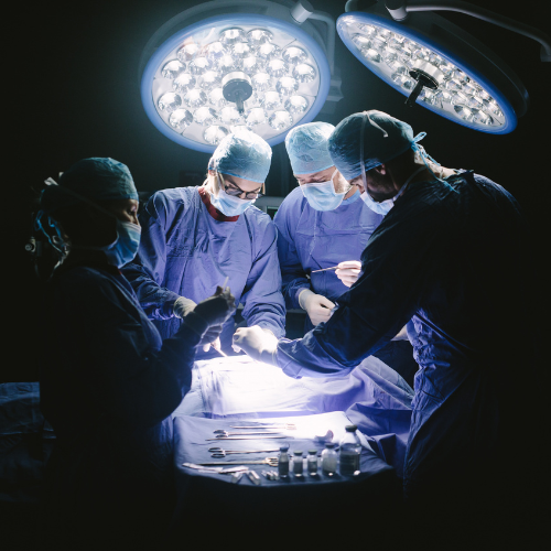 Equipe médica realizando uma cirurgia em centro cirúrgico, com quatro profissionais paramentados sob focos cirúrgicos e instrumentos organizados sobre a mesa ao lado do paciente.